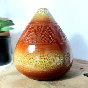Op de afbeelding: Een traanvormige keramische vaas met een glanzende afwerking, voorzien van een warm amberkleurig en goudgeel kleurverloop. Het bovenste gedeelte vertoont een gestructureerd kruisarceringpatroon, terwijl het onderste gedeelte overgaat in een gevlekt, poreus ogend geel glazuur.