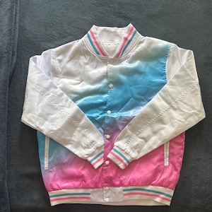 May include: A satin bomber jacket with a color gradient design. The jacket transitions from white to blue and then to pink. The collar and cuffs have striped detailing in pink and blue. The jacket has a button-up front.