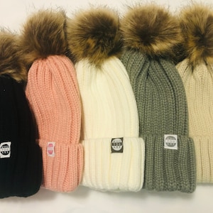 May include: Five knitted beanies with faux fur pom poms in black, pink, white, green, and beige. Each beanie has a small round patch with the text "WHO YOU ARE" in black and white.