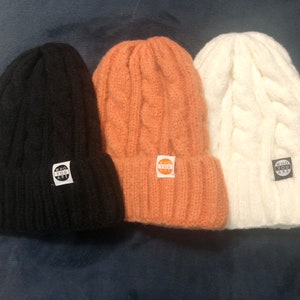 May include: Three knitted beanies in black, peach, and white with a cable knit pattern. Each beanie has a small round patch with the text "WHO YOU ARE" in white.