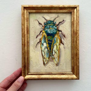 May include: Oil painting of a cicada in a gold frame. The insect is depicted with a teal and yellow body and wings, set against a cream-colored background. The frame has a detailed, ornate design.