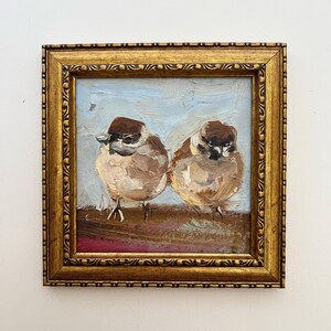 Puede incluir: Una pintura al &oacute;leo de dos peque&ntilde;os p&aacute;jaros marrones con marcas oscuras en la cabeza, de pie sobre una superficie marr&oacute;n contra un cielo azul claro. La obra est&aacute; encerrada en un marco cuadrado, ornamentado y dorado con relieve.