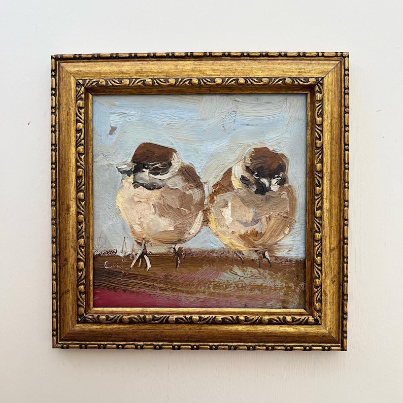Puede incluir: Una pintura al &oacute;leo de dos peque&ntilde;os p&aacute;jaros marrones con marcas oscuras en la cabeza, de pie sobre una superficie marr&oacute;n contra un cielo azul claro. La obra est&aacute; encerrada en un marco cuadrado, ornamentado y dorado con relieve.