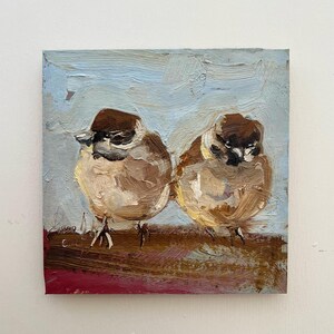 Puede incluir: Una pintura al &oacute;leo sobre lienzo cuadrado que muestra dos peque&ntilde;os p&aacute;jaros marrones y blancos posados uno al lado del otro. Los p&aacute;jaros tienen cabezas de color marr&oacute;n oscuro y cuerpos marrones claros, sobre un fondo azul claro texturizado.