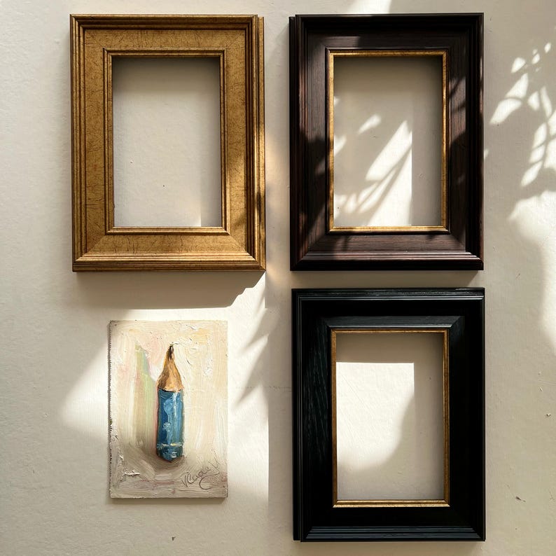 May include: Four rectangular picture frames in gold, dark brown, and black, with a small painting of a blue pencil. The frames are empty, with a gold inner border. The painting is on a small rectangular canvas.