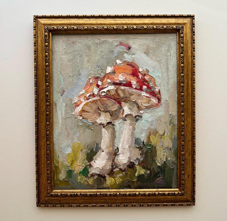 May include: An oil painting of two red and white mushrooms with thick, textured brushstrokes, set against a muted grey and green background. The artwork is enclosed in an ornate, rectangular gold-toned frame with a decorative carved border.