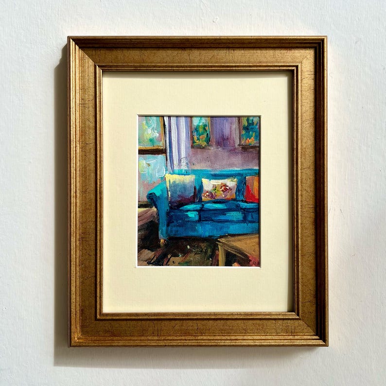 May include: An oil painting in a gold frame depicts a living room scene. A teal sofa with patterned pillows is the focal point. The background shows a window and artwork. The painting is framed with a cream-colored mat.