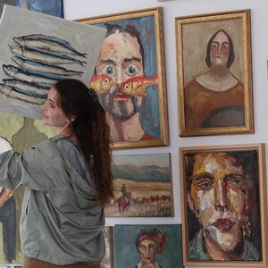 Puede incluir: Una mujer sostiene una pintura de peces frente a una pared que muestra varias pinturas al &oacute;leo enmarcadas. La obra de arte incluye retratos y paisajes, mostrando una diversa colecci&oacute;n de arte.