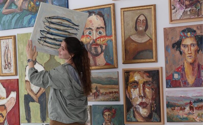 Puede incluir: Una mujer sostiene una pintura de peces frente a una pared que muestra varias pinturas al &oacute;leo enmarcadas. La obra de arte incluye retratos y paisajes, mostrando una diversa colecci&oacute;n de arte.