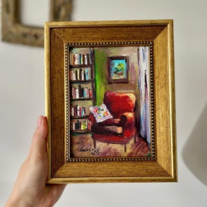 May include: A framed oil painting depicts a cozy reading nook. The artwork features a red armchair, a bookshelf, and a small framed picture. The painting is signed and dated. The frame is gold-colored.