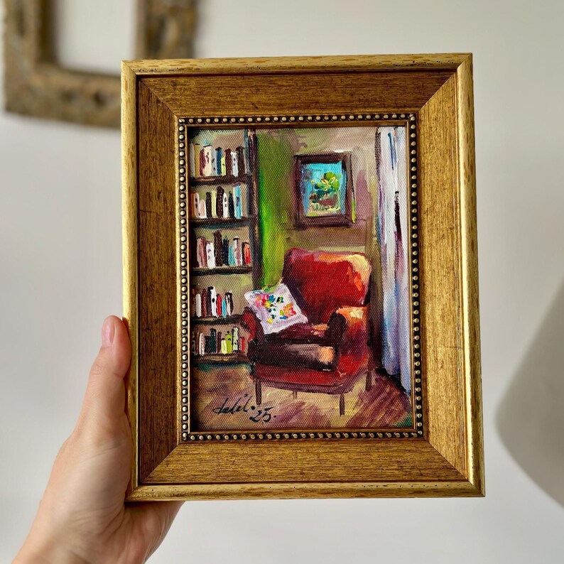 May include: A framed oil painting depicts a cozy reading nook. The artwork features a red armchair, a bookshelf, and a small framed picture. The painting is signed and dated. The frame is gold-colored.