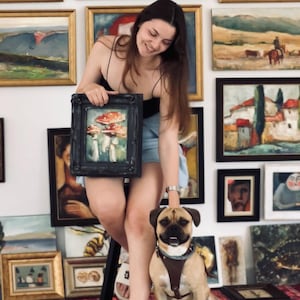 K&ouml;nnte beinhalten: Ein kleines gerahmtes &Ouml;lgem&auml;lde mit rot-wei&szlig;en Pilzen, das vor einer Wand mit verschiedenen gerahmten Landschafts- und Portr&auml;tbildern gehalten wird. Ein hellbrauner und schwarzer Hund mit braunem Ledergeschirr sitzt im Vordergrund.