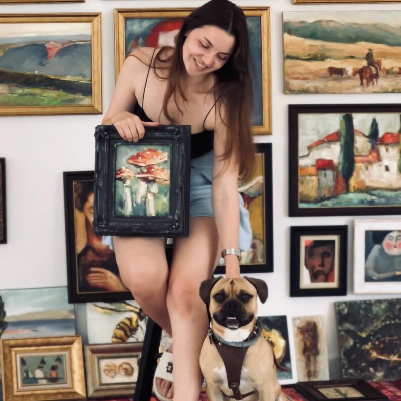 K&ouml;nnte beinhalten: Ein kleines gerahmtes &Ouml;lgem&auml;lde mit rot-wei&szlig;en Pilzen, das vor einer Wand mit verschiedenen gerahmten Landschafts- und Portr&auml;tbildern gehalten wird. Ein hellbrauner und schwarzer Hund mit braunem Ledergeschirr sitzt im Vordergrund.