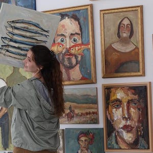May include: A woman holding a painting of fish in front of a wall displaying various framed oil paintings. The artwork includes portraits and landscapes, showcasing a diverse collection of art.