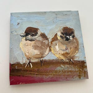 Puede incluir: Una pintura al &oacute;leo sobre lienzo cuadrado que muestra dos peque&ntilde;os p&aacute;jaros marrones y blancos posados uno al lado del otro. Los p&aacute;jaros tienen cabezas de color marr&oacute;n oscuro y plumaje texturizado, sobre un fondo de cielo azul claro.