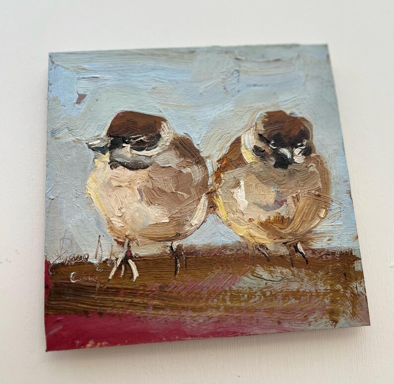 Puede incluir: Una pintura al &oacute;leo sobre lienzo cuadrado que muestra dos peque&ntilde;os p&aacute;jaros marrones y blancos posados uno al lado del otro. Los p&aacute;jaros tienen cabezas de color marr&oacute;n oscuro y plumaje texturizado, sobre un fondo de cielo azul claro.