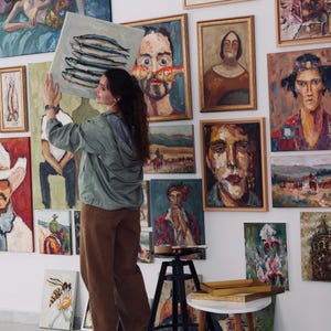 May include: An art gallery featuring numerous framed oil paintings. A person is holding a square canvas with a painting of fish. The paintings depict various subjects, including portraits, landscapes, and still lifes. The walls are white, and the floor is a light color.