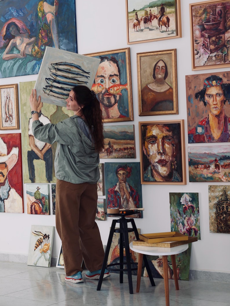 May include: An art gallery featuring numerous framed oil paintings. A person is holding a square canvas with a painting of fish. The paintings depict various subjects, including portraits, landscapes, and still lifes. The walls are white, and the floor is a light color.