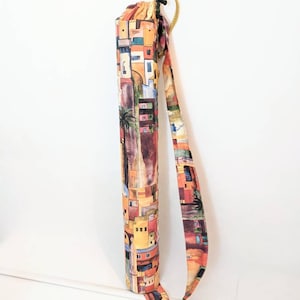 May include: A colorful fabric yoga mat bag with a long strap. The bag features a pattern of buildings and palm trees in shades of orange, yellow, brown, and green.