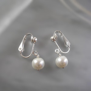 May include: A pair of clip-on earrings featuring a silver-toned metal design. Each earring has a small, round pearl pendant. The earrings are displayed against a soft, gray background.