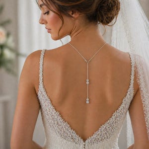 May include: A bride wearing a white wedding dress with a low back and beaded straps. A delicate silver necklace with three sparkling pendants hangs down the bride's back. A sheer veil drapes over her shoulders.