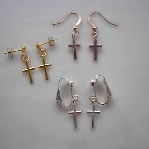 Könnte beinhalten: Eine Auswahl an Kreuzohrringen in Gold-, Silber- und Roségoldtönen. Die Ohrringe umfassen Stecker-, Haken- und Clip-on-Stile, jeweils mit einem einfachen Kreuzanhänger. Der Schmuck wird auf einer hellgrauen Oberfläche präsentiert.