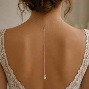 Op de afbeelding: Een zilveren rugketting met een fijne ketting en een enkele parel hanger. De ketting wordt gedragen over een witte kanten jurk, wat een elegante uitstraling creëert. De achterkant van de jurk is zichtbaar en toont de ingewikkelde kanten details.