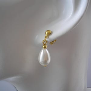 Puede incluir: Pendiente de clip dorado con un colgante de perla de imitaci&oacute;n blanca en forma de l&aacute;grima. El pendiente est&aacute; sujeto a un perno de color dorado que se engancha a la oreja. El pendiente se muestra sobre un fondo blanco.