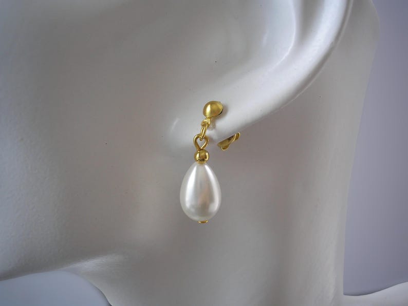 Puede incluir: Pendiente de clip dorado con un colgante de perla de imitaci&oacute;n blanca en forma de l&aacute;grima. El pendiente est&aacute; sujeto a un perno de color dorado que se engancha a la oreja. El pendiente se muestra sobre un fondo blanco.