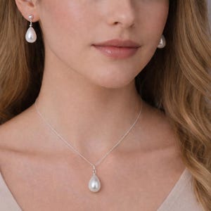 Puede incluir: Una mujer lleva un collar de perlas y pendientes a juego. La joyer&iacute;a presenta perlas en forma de l&aacute;grima suspendidas de delicadas cadenas de plata. Los pendientes cuelgan con gracia y el colgante del collar descansa sobre el pecho. Las perlas tienen un aspecto blanco y brillante.