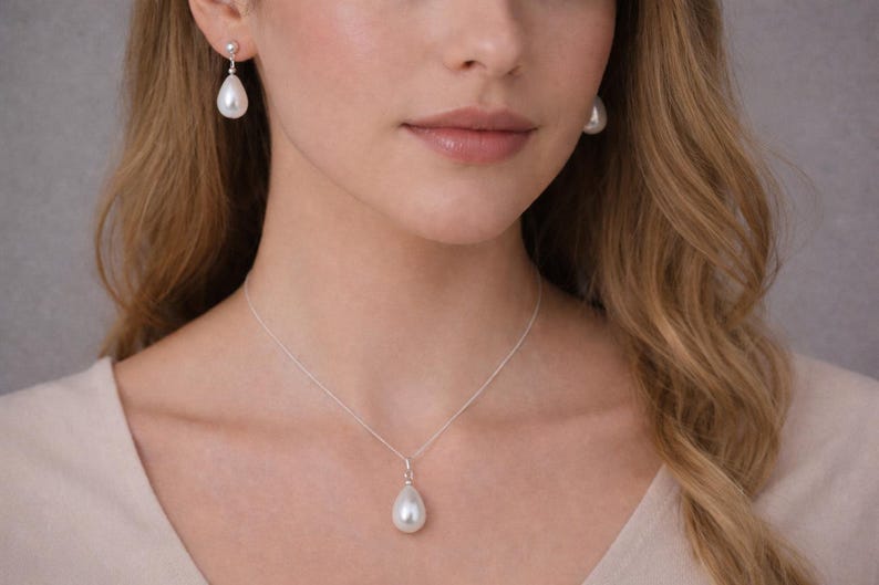 Puede incluir: Una mujer lleva un collar de perlas y pendientes a juego. La joyer&iacute;a presenta perlas en forma de l&aacute;grima suspendidas de delicadas cadenas de plata. Los pendientes cuelgan con gracia y el colgante del collar descansa sobre el pecho. Las perlas tienen un aspecto blanco y brillante.
