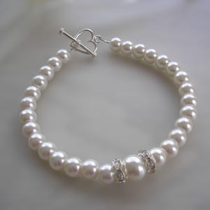 Könnte beinhalten: Ein weißes Perlenarmband mit einem herzförmigen Silberverschluss. Das Armband besteht aus einer Reihe runder, weißer Perlen. Zwei größere Perlen sind mit kleinen, klaren Kristallen verziert. Ein klassisches Schmuckstück.