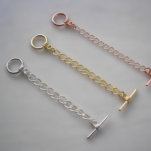Puede incluir: Tres cierres de palanca para joyería, cada uno con un anillo circular y una barra en T conectados por una cadena corta. El conjunto incluye un cierre de metal plateado, uno dorado y uno de color oro rosa, dispuestos en una formación diagonal.