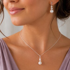 May include: A silver chain necklace featuring a pendant with two small clear beads and one larger white pearl, paired with matching drop earrings that each consist of two small clear beads and one larger white pearl on a silver post.
