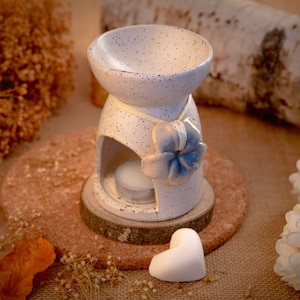 Op de afbeelding: Witte keramische olieverdamper met een blauw bloemenontwerp en een witte was smelt. De diffuser staat op een houten basis en een bruin oppervlak.