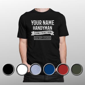 Puede incluir: Camiseta negra con el texto blanco "YOUR NAME HANDYMAN FIXING EVERYTHING EXCEPT STUPID". La camiseta tiene cuello redondo y mangas cortas. Las opciones de color se muestran debajo de la camiseta.