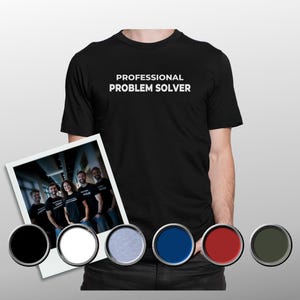 Op de afbeelding: Zwart T-shirt met de witte tekst "PROFESSIONAL PROBLEM SOLVER". De afbeelding toont ook een foto met een groep mensen en een selectie van kleurstalen.