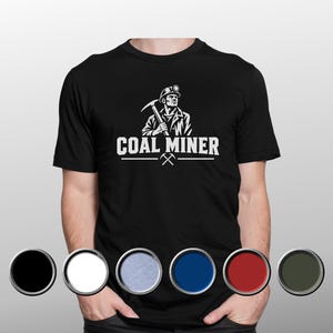 Könnte beinhalten: Ein schwarzes Kurzarm-T-Shirt mit einer weißen Grafik eines Bergmanns, der eine Spitzhacke hält, und dem darunter gedruckten Text COAL MINER. Unten sind sechs kreisförmige Farbmuster in Schwarz, Weiß, Hellblau, Blau, Rot und Dunkelgrün abgebildet.