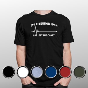 May include: A black short-sleeved t-shirt featuring white text that reads "MY ATTENTION SPAN HAS LEFT THE CHART" above a white heartbeat line graphic. Below the shirt are six circular colour swatches in black, white, light blue, blue, red, and dark green.
