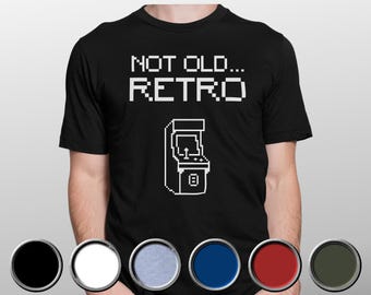 May include: A black short-sleeved t-shirt featuring white pixelated text that reads NOT OLD... RETRO above a white pixelated graphic of an arcade cabinet. Below the shirt are six circular color swatches in black, white, light blue, blue, red, and dark green.