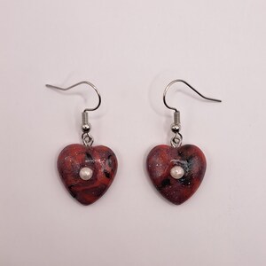 Könnte beinhalten: Ein Paar herzförmige Ohrringe. Die Ohrringe haben ein rot-schwarzes Marmormuster mit einer kleinen, runden, weißen Perle in der Mitte. Sie hängen an silberfarbenen Fischhaken-Ohrdrähten.