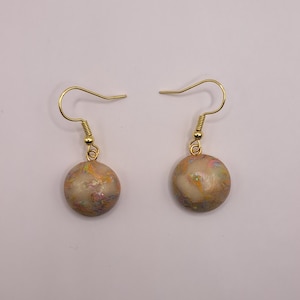 May include: A pair of handmade earrings with gold-colored hooks. Each earring features a round, beige bead with colorful glitter accents. The earrings are displayed against a plain, light background.