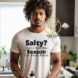 May include: A person wearing a white t-shirt with the text "Salty? That's How We Season Our Food, Baby!" and the logo "VIBE LIFE APPAREL". The person is holding a clear bottle with a white lid and a metal lid.