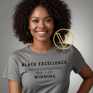May include: A gray t-shirt with black text that reads "BLACK EXCELLENCE It Ain't a Competition, But... I am WINNING". The shirt is being worn by a person with curly black hair. A gold logo with the text "VIBE LIFE APPAREL" is visible in the upper right corner of the image.