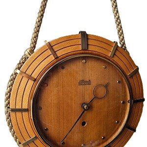 Puede incluir: Un reloj redondo de madera con una esfera marrón claro y un marco marrón oscuro. El reloj tiene una cubierta de vidrio transparente y manecillas y marcadores de color latón. Cuelga de un cordón trenzado de color dorado.
