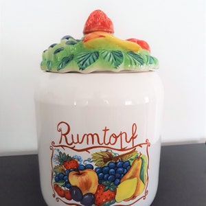 May include: White ceramic jar with a fruit design and a lid. The lid has a strawberry on top and the jar has a fruit design with the word "Rumtopf" written on it.