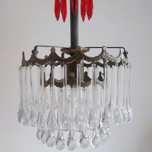 Op de afbeelding: Een vintage kroonluchter met een messing frame en heldere glazen druppelkristallen. De kroonluchter heeft een centrale fitting met vijf rode glazen druppelkristallen.