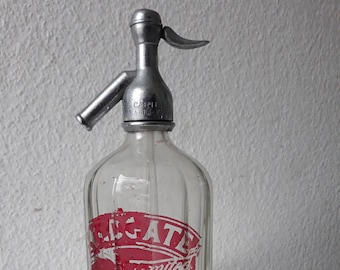 May include: A vintage glass bottle with a metal soda siphon top. The bottle has a red and white label with the text "LEGATE" and "mitco" on it.