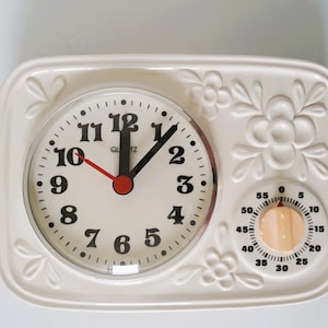 Puede incluir: Combinación de reloj y temporizador de cuarzo color crema con detalles florales en relieve. El reloj tiene números negros, manecillas negras y un segundero rojo. El temporizador tiene una perilla de madera y una esfera negra.