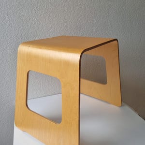 Könnte beinhalten: Ein hellfarbener Holzhocker mit einer glatten, geschwungenen Sitzfläche und zwei Seitenteilen, die jeweils einen großen, quadratischen Ausschnitt aufweisen. Der Hocker besteht aus gebogenem Sperrholz mit einer natürlichen Holzmaserung.